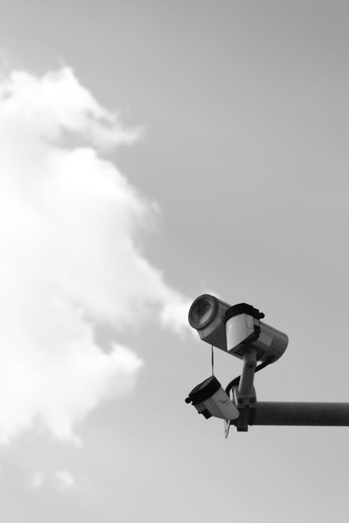 Black and white image of a surveillance camera mounted against a cloudy sky, representing security and monitoring.